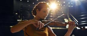 A woman playing the violin with a stage light shining over her shoulder and a concert band in the background.